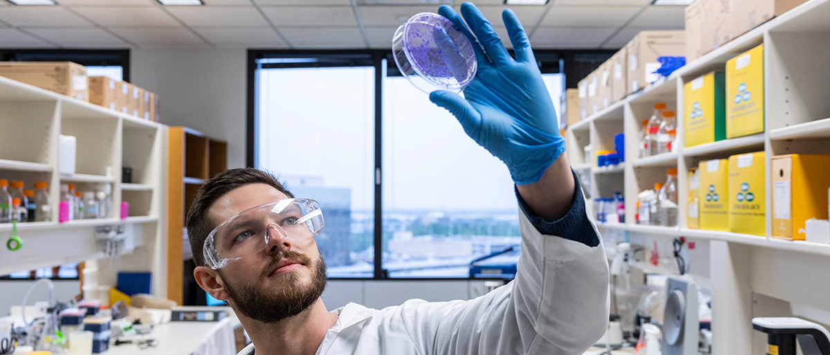 Man with dark hair and facial hair, wearing a white lab coat and goggles, examines purple substance in a petri dish.