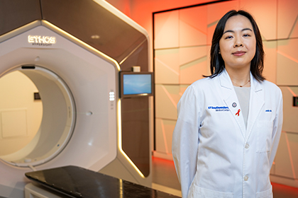 Smiling woman with dark hair wearing a white UT Southwestern Medical Center lab coat, standing next to a large Ethos imaging machine.