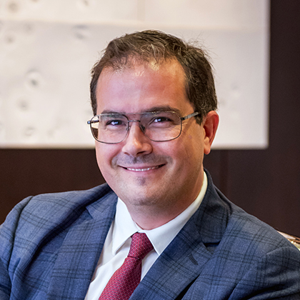 Smiling man with short dark hair and glasses, wearing a blue plaid suit, white shirt, and red tie.