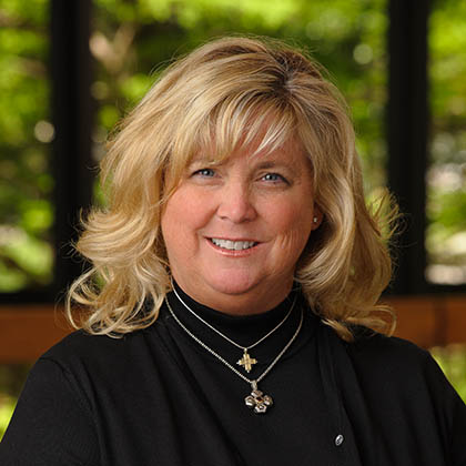 Smiling woman with long blond hair wearing a black top and 2 gold necklaces.