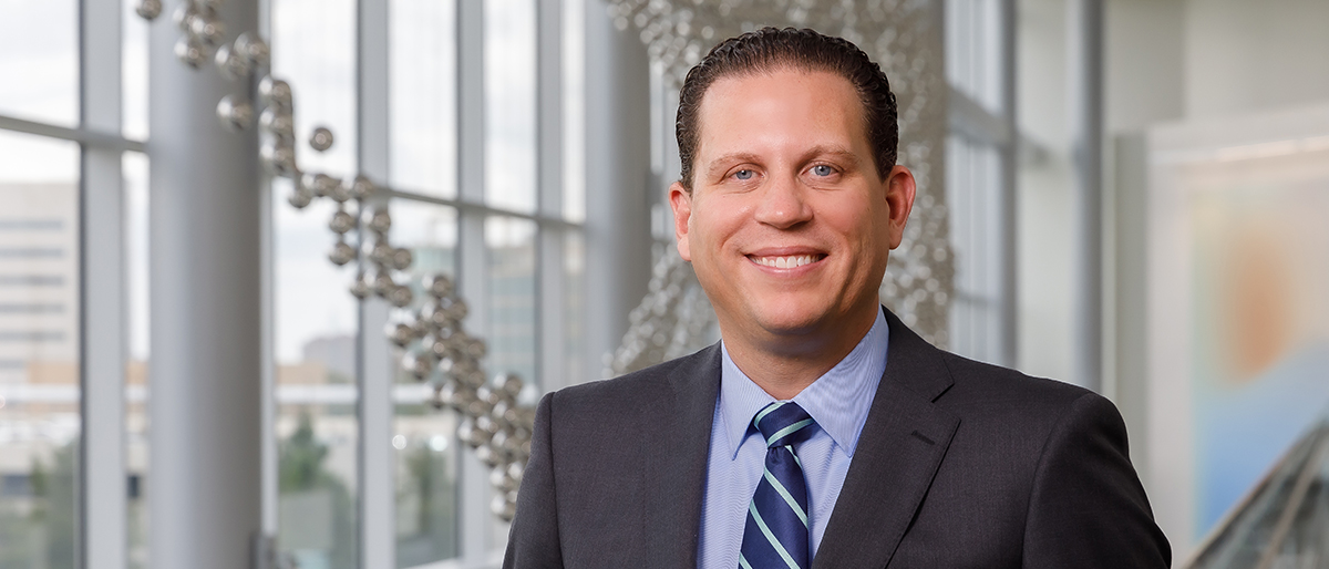 Smiling man with brown hair, wearing a dark gray suit, blue shirt, and blue striped tie.