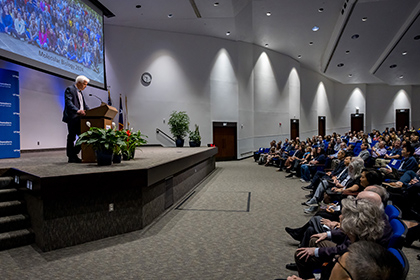 White-haired man wearing a gray suit, standing behind a podium on-stage, speaks to audience. Background on screen is a group photo of Molecular Biology 2024.