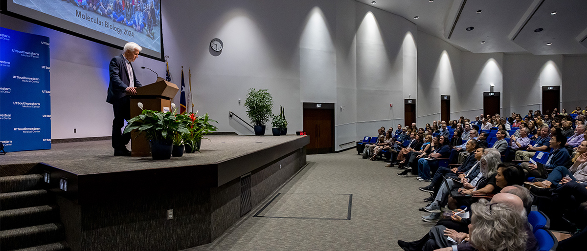White-haired man wearing a gray suit, standing behind a podium on-stage, speaks to audience. Background on screen is a group photo of Molecular Biology 2024.