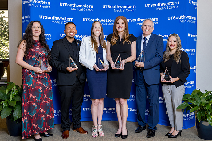 The 6 awardees stand in front of a blue UTSouthwestern Medical Center backdrop, holding their triangular glass awards.