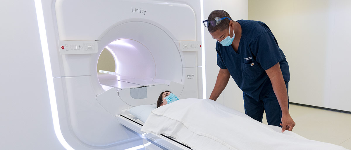 woman with dark hair and facemask lies under blanket on table going into Unity MRI machine with black male with mask and blue scrubs adjusts blanket