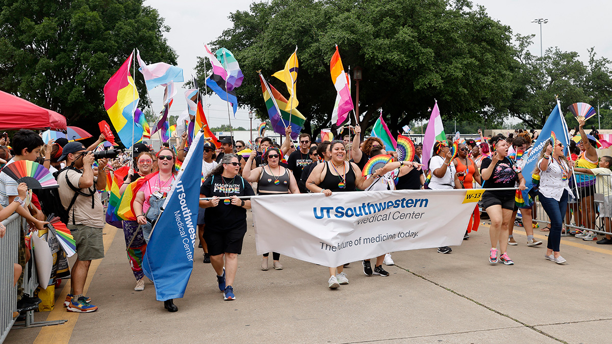 A group of men and women in summer attire parade down a street carrying multi-color flags and banners that read - UT Southwestern Medical Center, The Future of Medicine