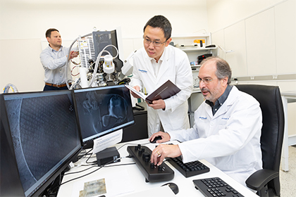 Two men wearing white lab coats, looking at scientific information on two computer monitors, while a man in the background works on machinery.