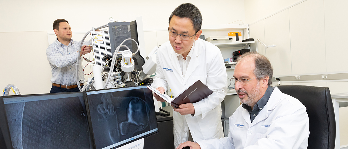 Two men wearing white lab coats, looking at scientific information on two computer monitors, while a man in the background works on machinery.