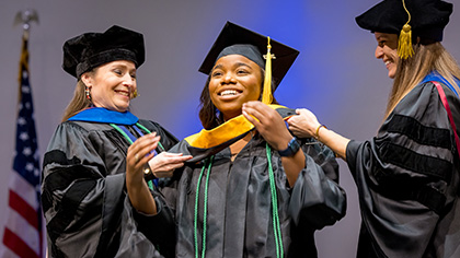3 women in cap and gowns placing gold sash on woman in center