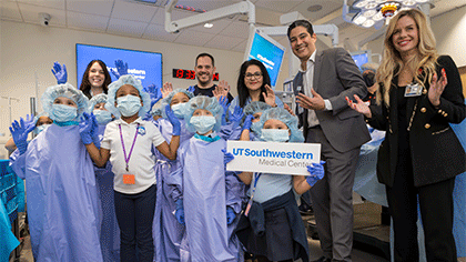 A group of children wearing scrubs, caps, masks, and gloves with a group of 5 adults, prepare to learn at the Simulation Center