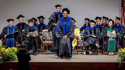 group of academics in cap and gown sit on stage behind podium; tall dark haired man with glasses stands behind smiling black woman graduate to adjust her sash