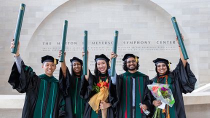 5 diverse medical school graduates in cap and gown smile and hold diploma tubes high