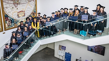 large group of graduates in cap/gown assemble on a white staircase with large map artwork behind on the wall