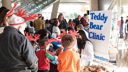 Adults and children wearing red foam reindeer antlers, watch young woman with long dark hear wearing PPE work on a teddy bear at a table with a sign that says, 'teddy bear clinic ut southwestern medical center' with other people standing around in the background