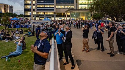 crowd of people on lawn outside hospital, all wearing eclipse glasses and gazing at the sky