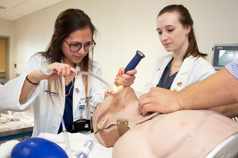 In 2019, a group of medical students practice clinical procedures. Training is heavily focused on working as part of medical care teams. Woman in lab coat holding light and pushing tube down dummy's throat