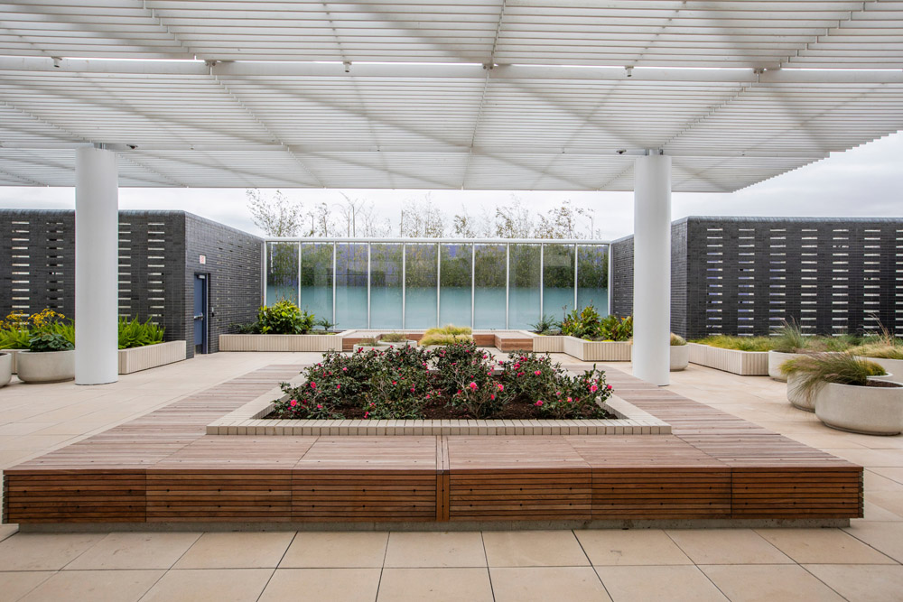 The psychiatric unit features 5,000 square feet of outdoor space and a wellness garden designed to provide a positive-care environment and recreation space for patients. Courtyard with flowers, wood seating, and shaded cover