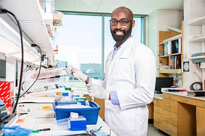 Smiling bald man with glasses and dark facial hair in a lab, wearing a white lab coat, holding a dropper and test tube.