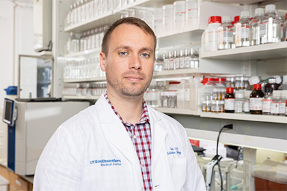 Man with brown hair and facial scruff in a lab, wearing a white lab coat over a red plaid shirt.