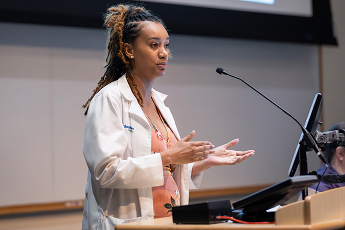 Dark-haired woman in lab coat speaking at podium.
