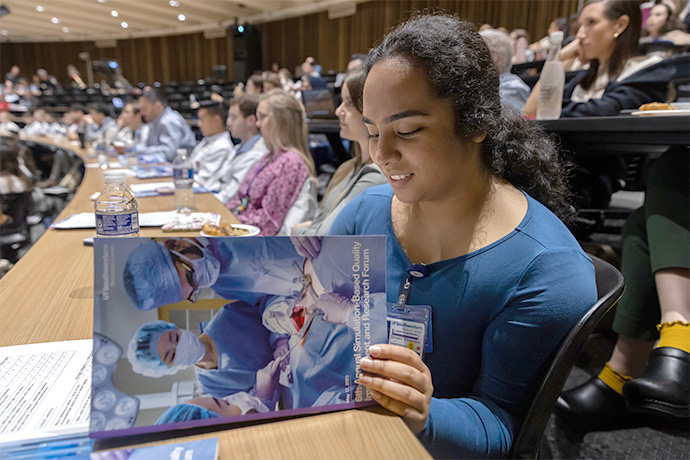 Woman looking at book in forground, audience seated at rows of tables in background watching lecture.