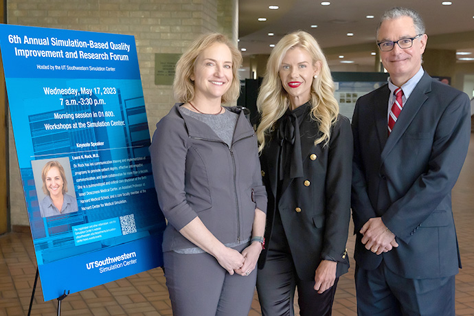 Left to right, blue poster, blond woman in gray jacket, blond woman in black suit, man with glasses wearing dark suit.