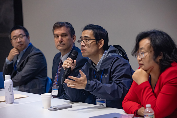 AI Symposium audience Three men and one woman seated at a table. One man is speaking emphatically into a microphone.