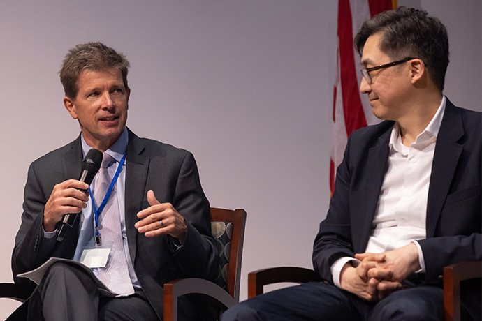 AI Symposium Man with short brown hair, wearing a gray suit, speaking into a microphone, and glancing at a man with dark hair and glasses wearing a dark suit seated next to him.