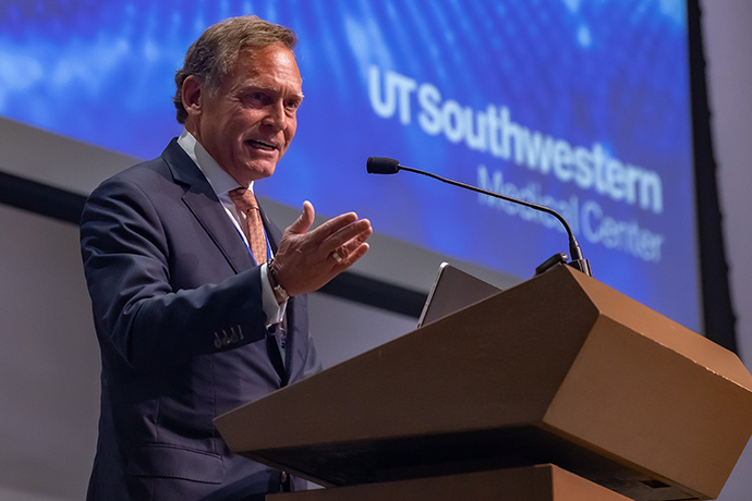 AI Symposium Man with dark, graying hair, wearing a gray suit and glasses, gesturing with his hands as he speaks from a podium. UT Southwestern Medical Center logo is displayed on the screen behind him.