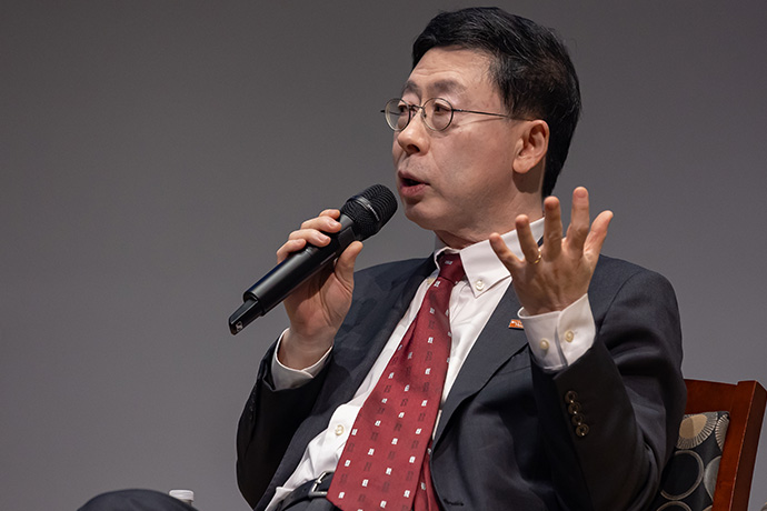 AI Symposium Seated man with dark hair wearing a gray suit, white shirt, red tie, and wire-rimmed glasses speaks into a microphone and gestures with his hands.