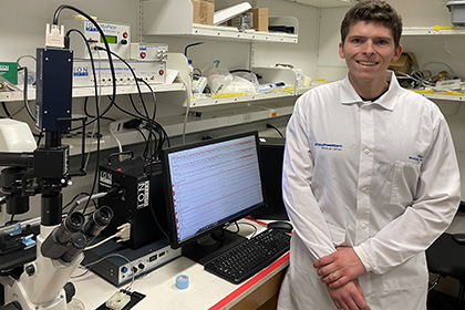 Smiling man with curly brown hair wearing a white lab coat, standing in a lab.