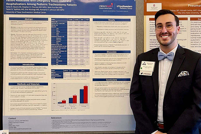 Smiling man with dark hair, trim beard, and mustache, wearing a dark suit and tie, standing next to poster of his ressearch on Factors Associated with Emergency room visits and hospitilizations among pediatric tracheostomy patients for Chilren's Health and UT Southwestern Pediatric group.