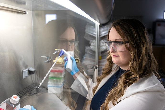 Looking in glass Woman with long brown highlighted hair, wearing a white lab coat over a dark top and protective glasses, holding a large dropper over a small white container. Her image is reflected in glass protecting the samples.