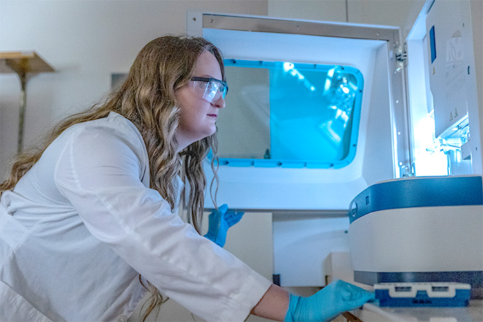 Woman with long brown highlighted hair, wearing a white lab coat and protective glasses, leans over to look at a piece of lab equipment.
