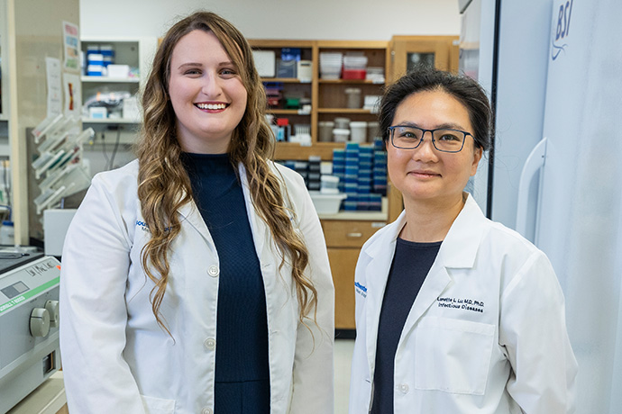 Scholar with mentor Two smiling women wearing white lab coats, posed in a lab. Woman, right, lab coat reads-Lennette L. Lu, M.D. Ph.D., Infectious Diseases.
