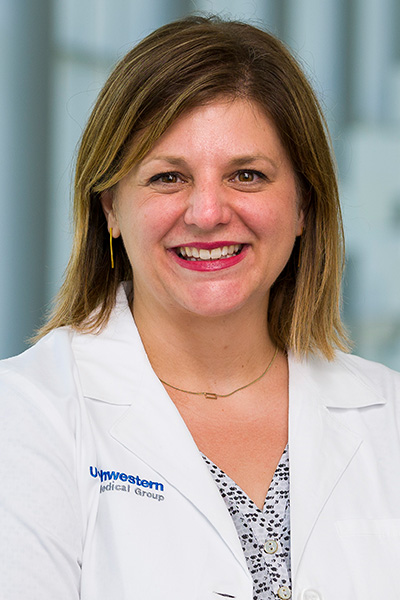 Smiling woman with highlighted brown hair wearing a white UT Southwestern Medical Center lab coat over a black, white, and gray blouse.