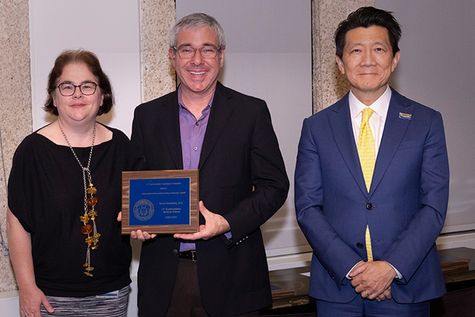 One woman and two men wearing dress clothes. Man in the center is holding an unreadable blue plaque on a wood background.