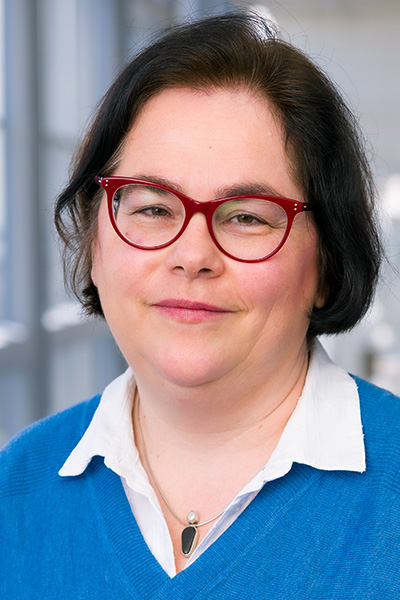 Smiling woman with short dark hair wearing a blue sweater over a white blouse, a decorative necklace and red-framed glasses.