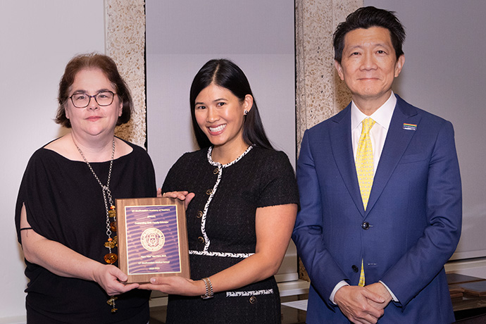 Two women wearing dress clothes holding a blue unreadable plaque on a wood background, standing with a man in a blue suit.