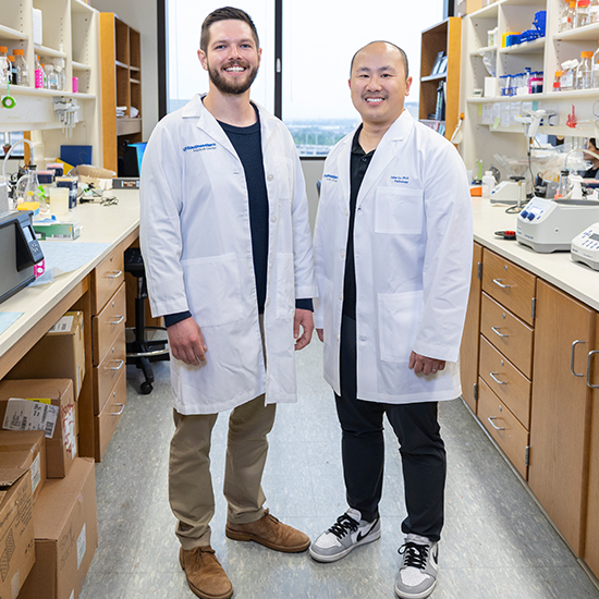 Two smiling men wearing white lab coats, standing in a medical lab.