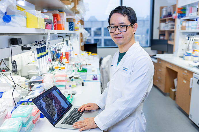 Smiling man wearing a white lab coat, standing in a lab in front of an open laptop computer.