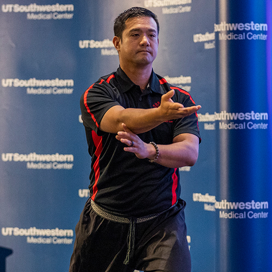 Man with dark hair, wearing black and red athletic cloths, stands with one arm outstretched. Background, UT Southwestern Medical Center.