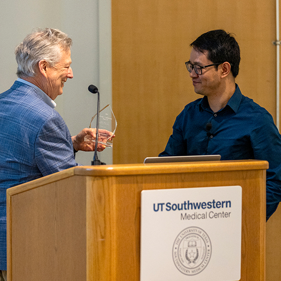 Two men standing behind a UT Southwestern medical podium, one handing the other a tear-drop shapped glass award.