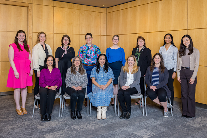 Thirteen women posing as a roup in front of a paneled wall.