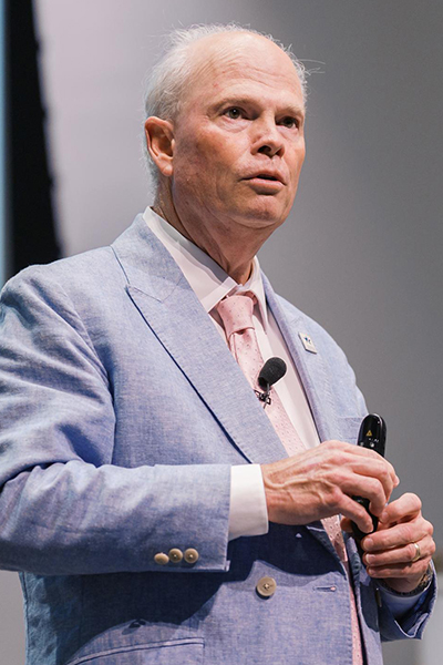Man with receding white hair, wearing a blue jacket, white shirt, and pink tie, holding a remote control.