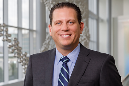 Smiling man with brown hair, wearing a dark gray suit, blue shirt, and blue striped tie.