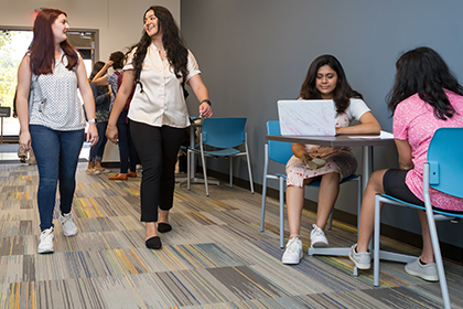 2 young women walking in hallway near more students seated at tables along the wall
