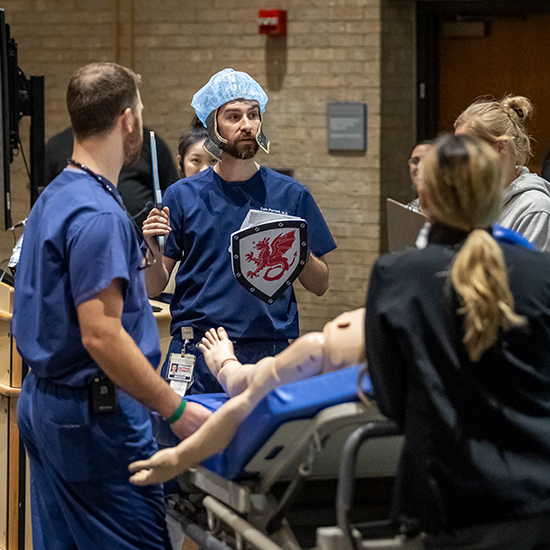 Dr. Samuel Parnell wears blue scrubs with fake armor helmet and shield standing at foot of bed with simulated patient as several residents watch