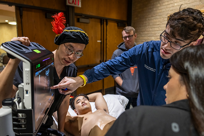 Dr. Zhao wearing feathered cap observes 3 medical residents looking at monitor as simulation center patient lies on exam table