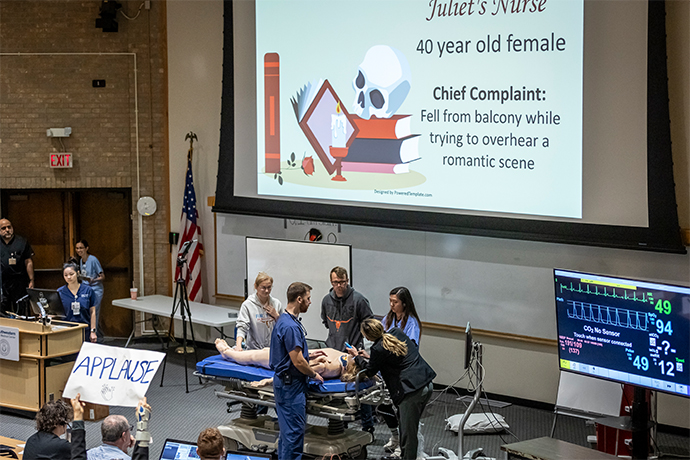 residents in lecture hall gather under presentation screen titled Juliet's Nurse; 5 stand around simulated patient on gurney, large display shows vital signs (right) as others watch in audience
